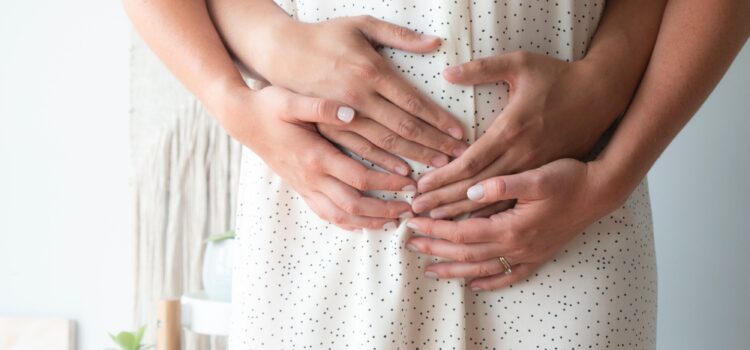 Photo focuses on the mid section of a woman in early pregnancy. Two hands reach around from the back to hold her belly. Her hands are placed on top of theirs.