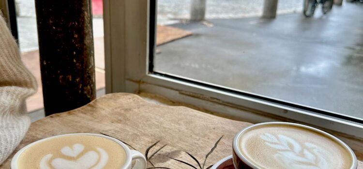 Two lattes in cups on saucers, side by side on a wooden table in a café in Paris with a view out the window of a rainy day