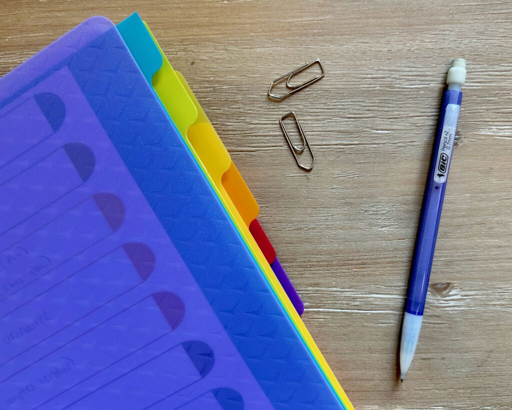 colored divider tabs sticking out of semi transparent binder with two paperclips and a mechanical pencil off to the side