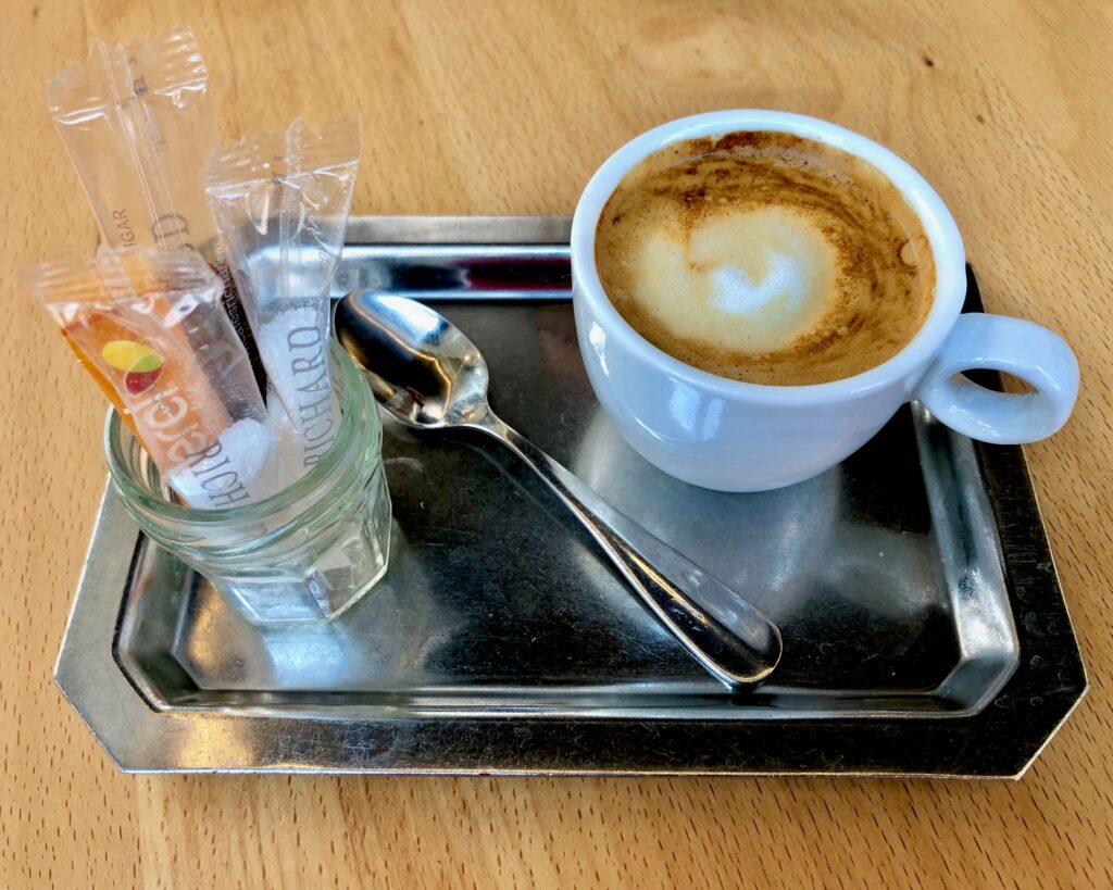 Un café noisette (an espresso with a splash of milk) in a small white espresso cup. It's on a rectangular silver tray with a silver spoon along with a small jar filled with plastic packets of sugar.