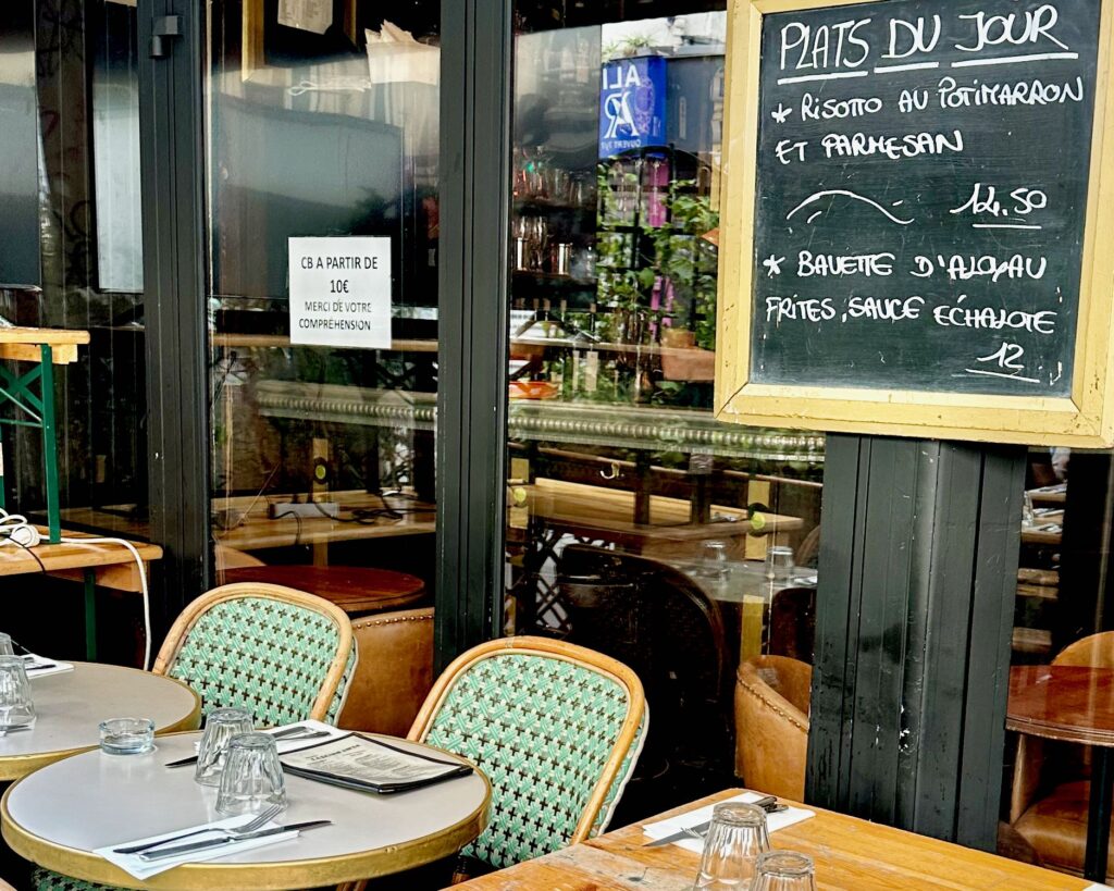 Typed sign reading, "CB A PARTIR DE 10€ MERCI DE VOTRE COMPREHENSION." This sign is outside of a Parisian café restaurant to indicate that they only accept credit card payments with a minimum 10 euro purchase. Black board listing the daily specials hanging up. Tables on the terrasse are set for meal service with forks, knives, napkins, and water glasses.