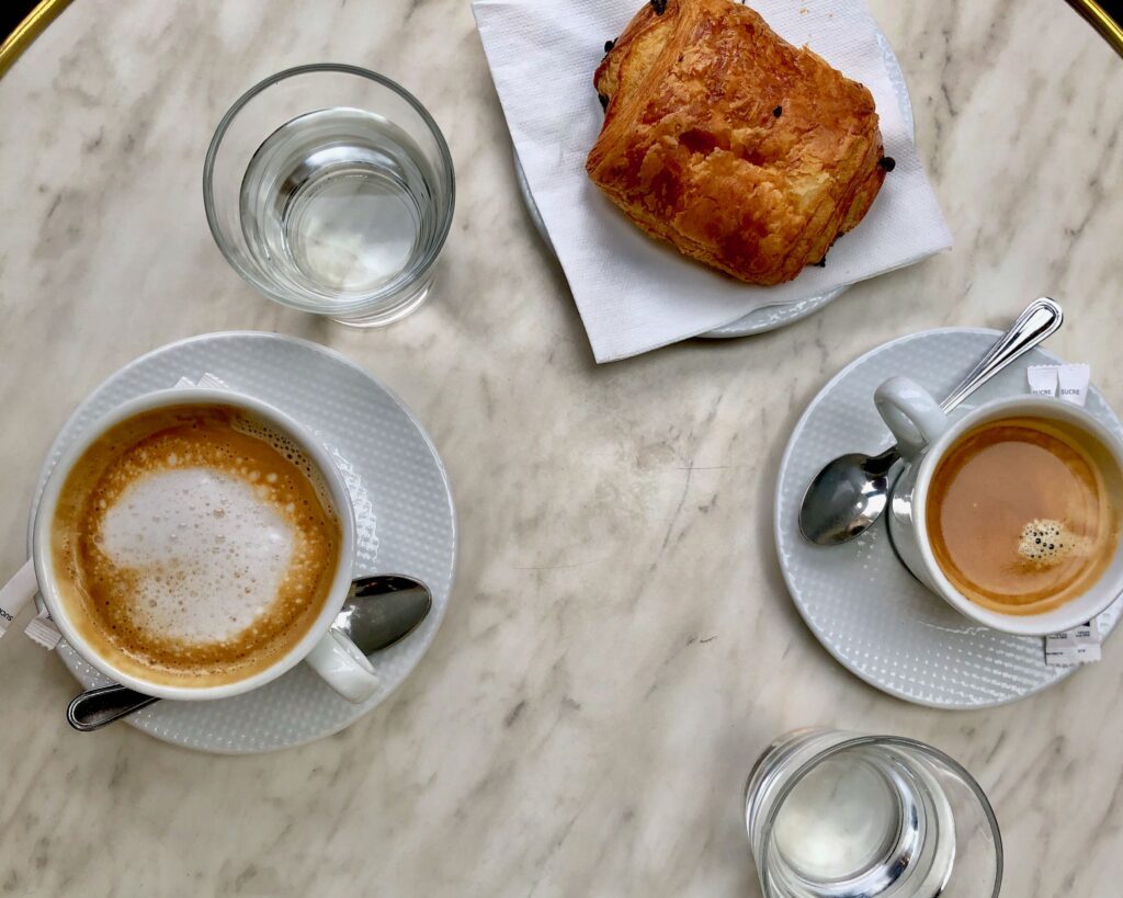 aerial view of two coffees, two glasses of water, and a pain au chocolat on a white marble table on a terrace in Paris