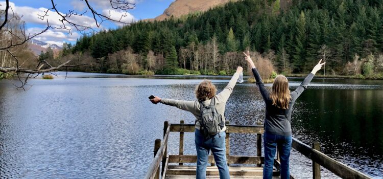 3 Things Americans Abroad Should Do Every Year Two American women abroad in Scotland, visiting Glenco Lochan, facing the lake with backs to the camera