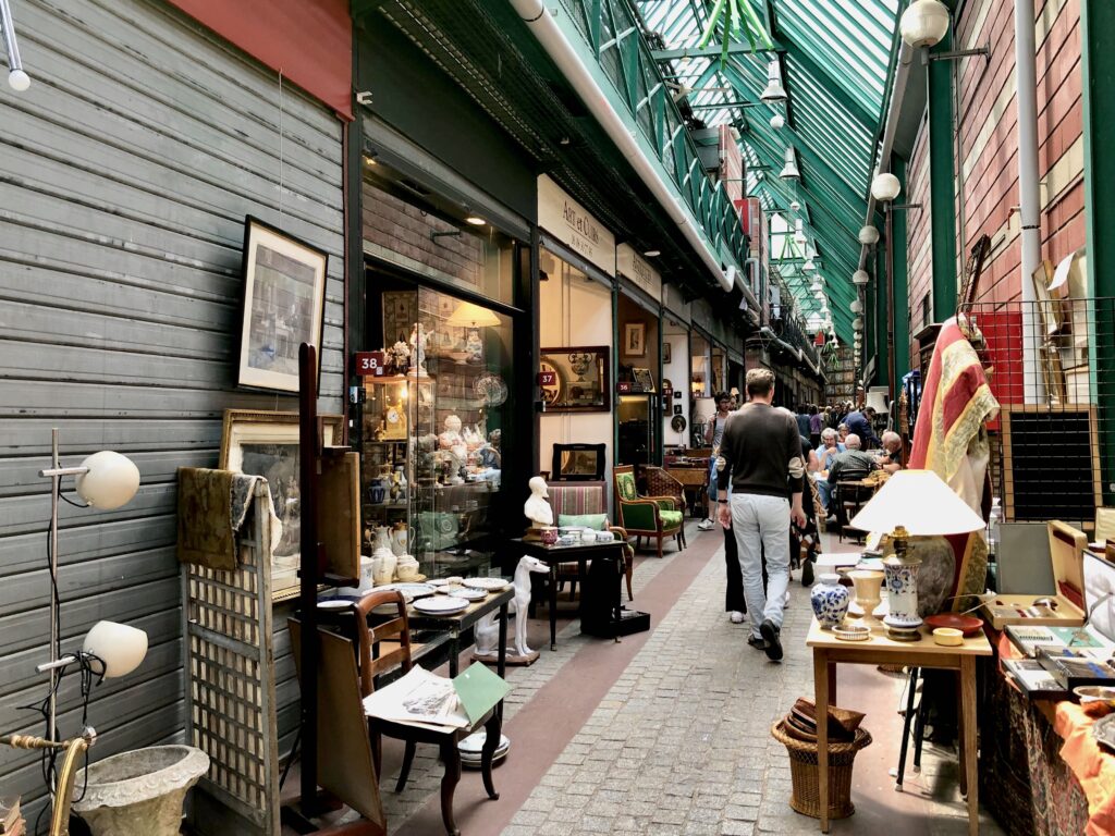 Inside the marché dauphine, a glass-ceilinged pavillion and one of the covered flea markets at Paris Saint-Ouen. Antiques are lined up along both sides of the alleyway.