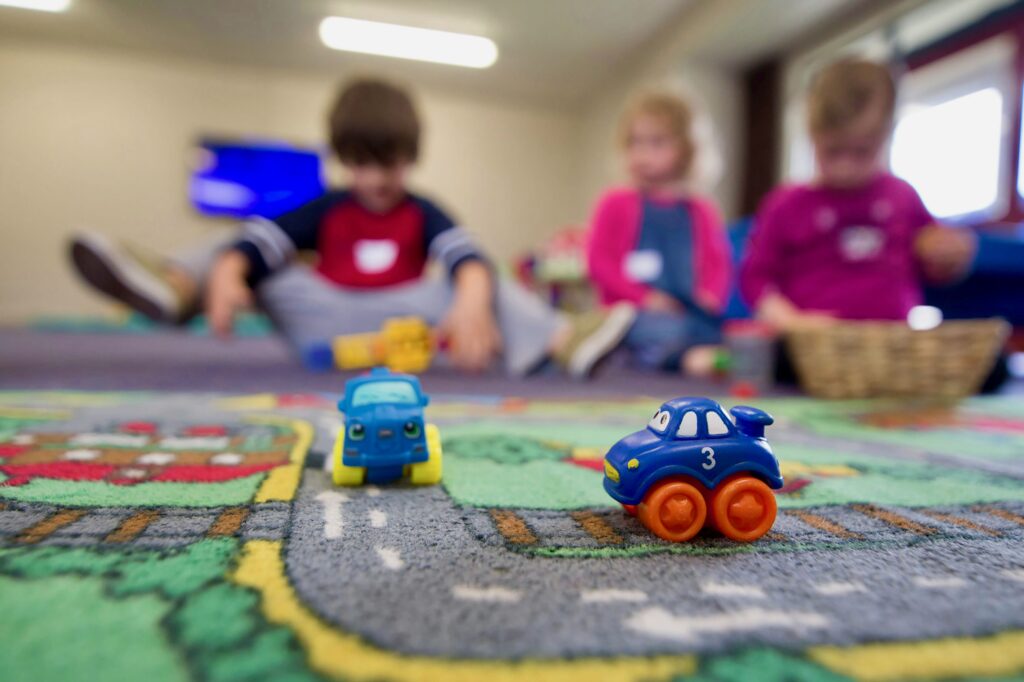 Two small plastic cars on a child's play rug with three kids sitting on the floor in the background