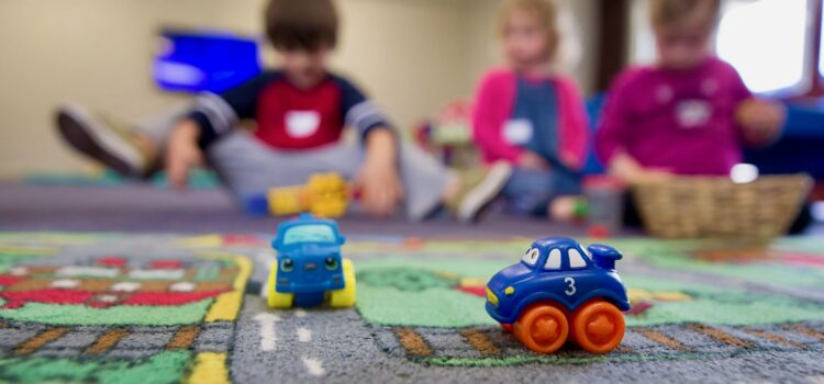 Two small plastic cars on a child's play rug with three kids sitting on the floor in the background