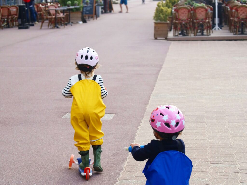 Two toddlers moving away from the camera on kick scooters on a sidewalk in France
