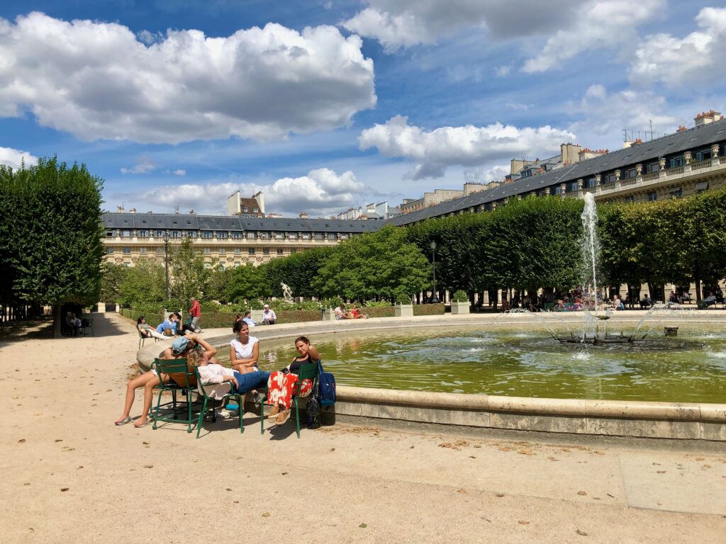 A group of women chatting and sitting in iconic green chairs near a fountain in the Jardin du Palais Royal in Paris.